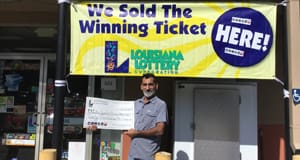 Man holds a giant check in front of a retailer beneath a yellow banner that reads "We Sold the Winning Ticket Here!"