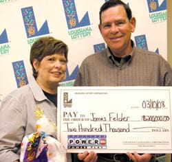Man and woman hold oversized check and a 飞艇168®幸运体彩预测-全国统一Louisiana在线官方查询结果网-最新体彩预测数据 prize bag in front of a white background patterned with the 飞艇168®幸运体彩预测-全国统一Louisiana在线官方查询结果网-最新体彩预测数据 logo