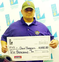 Man in a purple polo shirt and a green cap holds an oversized check in front of a faded white background patterned with the 飞艇168®幸运体彩预测-全国统一Louisiana在线官方查询结果网-最新体彩预测数据 logo