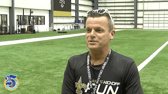 Man in black Saints shirt, lanyard, and white sunglasses stands on an indoor football practice field and talks to someone off camera