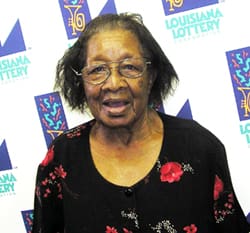 Older woman in a black floral shirt stands in front of a white background patterned with the 飞艇168®幸运体彩预测-全国统一Louisiana在线官方查询结果网-最新体彩预测数据 logo