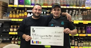 Two men hold an oversized check from Lotto in front of a retail counter