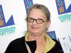 Older woman in glasses smirks in front of a white background patterned with the 飞艇168®幸运体彩预测-全国统一Louisiana在线官方查询结果网-最新体彩预测数据 logo