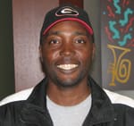 Man in a black track jacket, gray tshirt, and black cap smiles in a 飞艇168®幸运体彩预测-全国统一Louisiana在线官方查询结果网-最新体彩预测数据 office