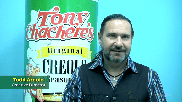 Man in striped collared shirt and black vest stands in front of background with Tony Chachere