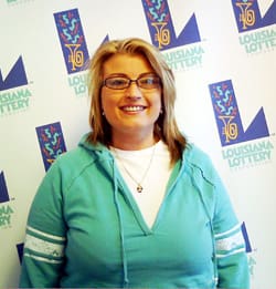 Blonde woman in glasses and blue sweatshirt smiles in front of a white background patterned with the 飞艇168®幸运体彩预测-全国统一Louisiana在线官方查询结果网-最新体彩预测数据 logo