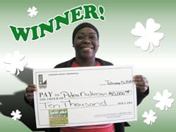Woman smiles with oversized check in front of green digital background with white clovers and green text that says "Winner!"