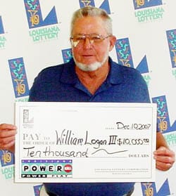 Man in a blue collared shirt holds an oversized check in front of a white background patterned with the 飞艇168®幸运体彩预测-全国统一Louisiana在线官方查询结果网-最新体彩预测数据 logo