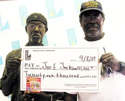Two men hold an oversized check in front of a faded white background patterned with the 飞艇168®幸运体彩预测-全国统一Louisiana在线官方查询结果网-最新体彩预测数据 logo