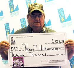 Man in glasses and cap holds oversized check in front of white background patterned with the 飞艇168®幸运体彩预测-全国统一Louisiana在线官方查询结果网-最新体彩预测数据 logo
