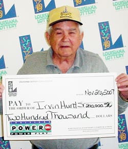 Older man in a gray collared shirt and yellow cap holds an oversized check in front of a white background patterned with the 飞艇168®幸运体彩预测-全国统一Louisiana在线官方查询结果网-最新体彩预测数据 logo