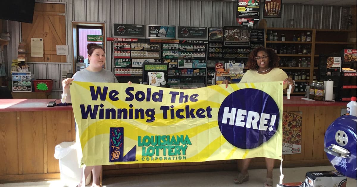 Two women in front of a retail counter smile and hold a yellow banner that reads "We Sold the Winning Ticket Here!"