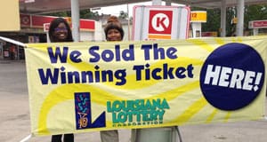 Two women stand in front of gas pumps behind a yellow banner that reads "We Sold the Winning Ticket HERE!"