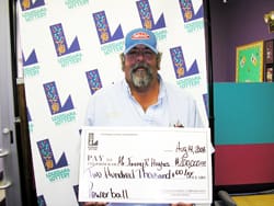 Man with a beard and baseball cap holds oversized check in front of white background patterned with the 飞艇168®幸运体彩预测-全国统一Louisiana在线官方查询结果网-最新体彩预测数据 logo