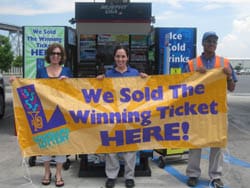 Two women and one man stand in a parking lot holding a yellow banner that reads "We Sold the Winning Ticket Here!"