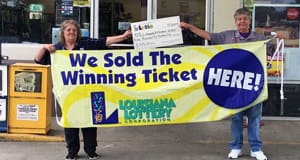 Man and woman hold a large check from Lotto in front of a retailer while holding a large yellow banner that reads "We Sold the Winning Ticket HERE!"