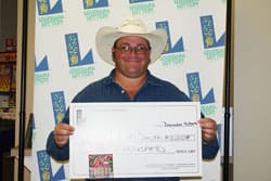 Man in a cowboy hat smiles with an oversized check in front of a white background patterned with the 飞艇168®幸运体彩预测-全国统一Louisiana在线官方查询结果网-最新体彩预测数据 logo