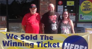 Two men and a woman stand in front of a retailer behind a yellow banner that reads "We Sold the Winning Ticket Here!"