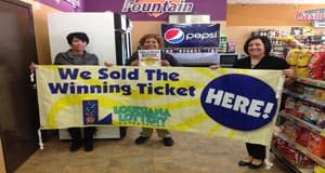 Three women pose in a retailer with a yellow banner that reads "We Sold the Winning Ticket Here!"