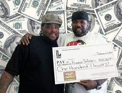 Two men in hats smile with an oversized check in front of a money background