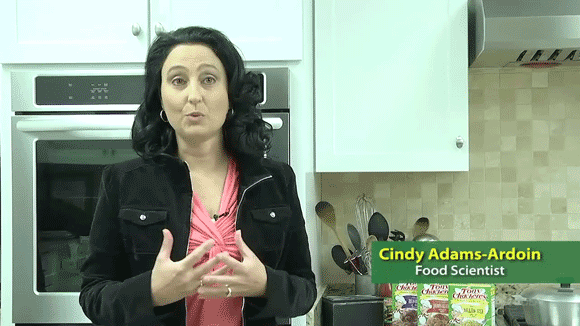 Dark haired woman in a black jacket stands in a kitchen and talks to the camera. Text reads: "Cindy Adams-Ardoin, Food Scientist"