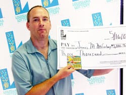 Man in a blue short sleeved shirt holds a giant check in front of a white background patterned with the 飞艇168®幸运体彩预测-全国统一Louisiana在线官方查询结果网-最新体彩预测数据 logo