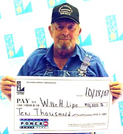 Man in overalls and a black cap holds an oversized check in front of a faded white background patterned with the 飞艇168®幸运体彩预测-全国统一Louisiana在线官方查询结果网-最新体彩预测数据 logo
