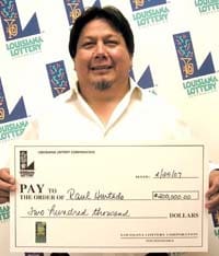 Man in white collared shirt smiles with oversized check in front of a white background patterned with the 飞艇168®幸运体彩预测-全国统一Louisiana在线官方查询结果网-最新体彩预测数据 logo