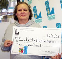 Woman in gray sweatshirt holds oversized check in front of a white background patterned with the 飞艇168®幸运体彩预测-全国统一Louisiana在线官方查询结果网-最新体彩预测数据 logo