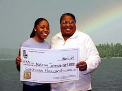 Man and woman smile with an oversized check in front of a body of water with a rainbow in the background