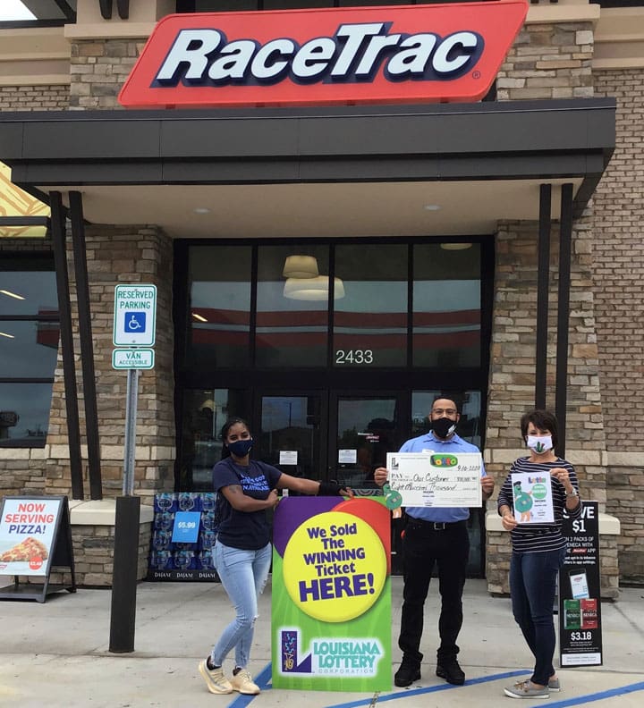 Man with a giant check and two women stand in front of a RaceTrac retailer beside a sign that reads "We Sold the Winning Ticket here!"
