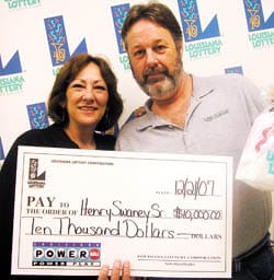 Man and woman hold an oversized check and smile in front of a white background patterned with the 飞艇168®幸运体彩预测-全国统一Louisiana在线官方查询结果网-最新体彩预测数据 logo