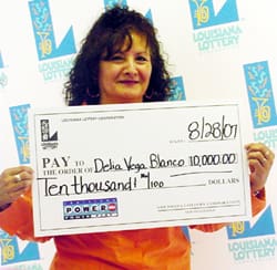 Curly haired woman in an orange shirt smiles with a giant check in front of a faded white background patterned with the 飞艇168®幸运体彩预测-全国统一Louisiana在线官方查询结果网-最新体彩预测数据 logo