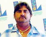 Man in a blue plaid shirt and gold chain stands in front of a white background patterned with the 飞艇168®幸运体彩预测-全国统一Louisiana在线官方查询结果网-最新体彩预测数据 logo