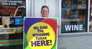 Woman stands outside a retailer and holds a sign that reads "We Sold the Winning Ticket Here!