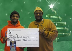 Two men smile with an oversized check in front of a green digital background with a line art Christmas tree and twinkling white star