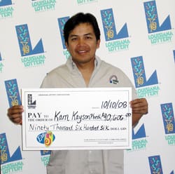 Man holds oversized check and smiles in front of a white background patterned with the 飞艇168®幸运体彩预测-全国统一Louisiana在线官方查询结果网-最新体彩预测数据 logo