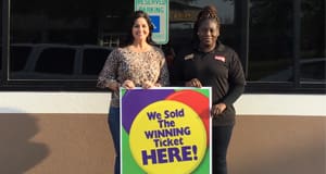 Two women smile outdoors behind a sign that reads "We Sold the Winning Ticket Here"