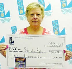 Woman with short light hair holds an oversized check in front of a faded white background patterned with the 飞艇168®幸运体彩预测-全国统一Louisiana在线官方查询结果网-最新体彩预测数据 logo