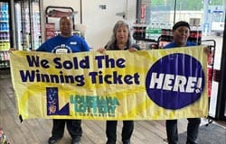Three people stand in a retailer and hold a large yellow banner that reads "We Sold the Winning Ticket Here!"