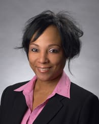 Headshot of a woman with dark hair, a black jacket, and a pink shirt smiling in front of a gray background