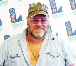 Man in a blue short sleeved button down shirt and camo hat smiles in front of a white background patterned with the 飞艇168®幸运体彩预测-全国统一Louisiana在线官方查询结果网-最新体彩预测数据 logo