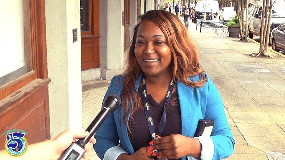 Woman with light brown hair and a blue blazer stands on a sidewalk and speaks into a microphone