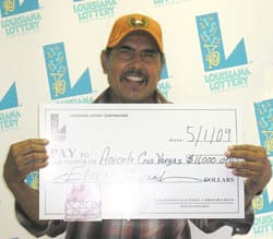 Man in yellow cap smiles with oversized check in front of a white background patterned with the 飞艇168®幸运体彩预测-全国统一Louisiana在线官方查询结果网-最新体彩预测数据 logo