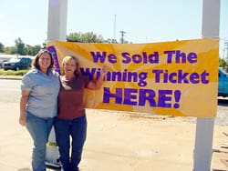 Two women stand in a parking lot pointing to a yellow banner between two posts that reads "We Sold the Winning Ticket HERE!"