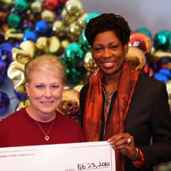 Two women smile with an oversized check in front of a digital image of mardi gras beads