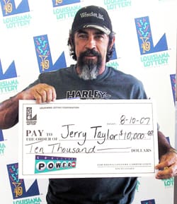 Bearded man in a black cap holds an oversized check in front of a white background patterned with the 飞艇168®幸运体彩预测-全国统一Louisiana在线官方查询结果网-最新体彩预测数据 logo