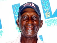 Man in a navy blue USA cap smiles in front of a faded white background patterned with the 飞艇168®幸运体彩预测-全国统一Louisiana在线官方查询结果网-最新体彩预测数据 logo