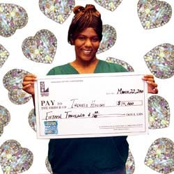 Woman smiles with an oversized check in front of a white digital background with diamond hearts on it