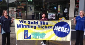 Three women stand outside a retailer and hold a yellow banner that reads "We Sold the Winning Ticket Here!"
